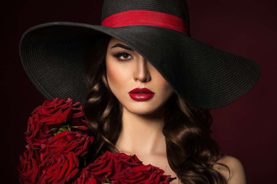 Portrait of a beautiful woman in a black hat with red roses on the shoulders. Red Lips. Beautiful Hair.